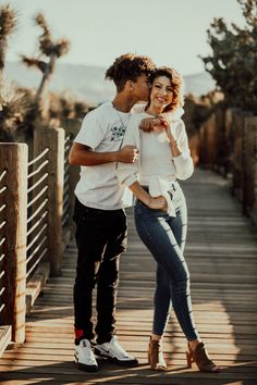 a man and woman standing on a wooden bridge with their arms around each other as they kiss