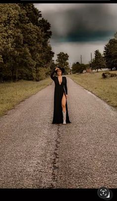 a woman in a long black dress standing on the side of a road with trees