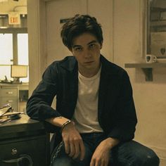 a young man sitting on top of a wooden table next to a black drawer and clock