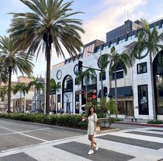 a woman crossing the street in front of a building with palm trees on both sides