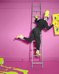 a man in black jumpsuit standing on a ladder next to a pink wall and painting