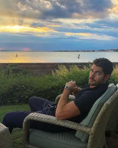 a man is sitting in a chair on the grass by the water with his arms crossed