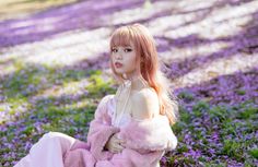 a young woman sitting on the ground in front of purple flowers wearing a pink fur coat