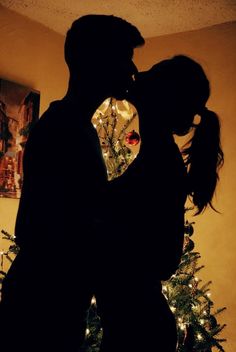 a man and woman kissing in front of a christmas tree with the lights on it