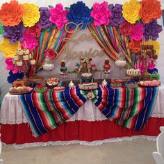 a table with colorful paper flowers and desserts on it's sides, in front of a backdrop