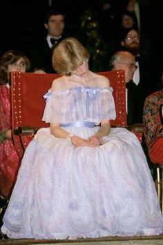 a woman in a white dress sitting on a red chair with other people behind her