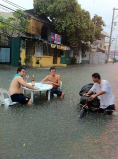 three men sitting at a table in the middle of a flooded street, one drinking beer