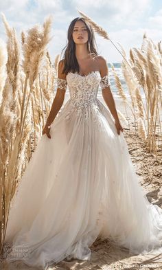 a woman in a white wedding dress standing on the beach with tall grass behind her