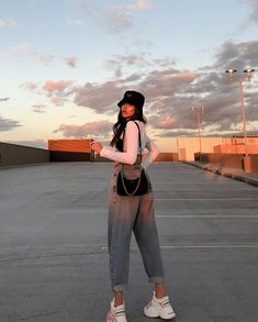 a woman standing in an empty parking lot with her hand on her hip and wearing a black hat