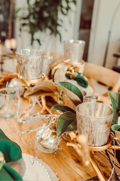 a wooden table topped with lots of glasses and plates covered in greenery next to candles