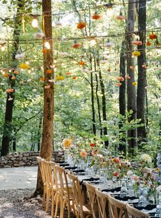 a long table set up for an outdoor dinner in the woods with flowers and hanging lights