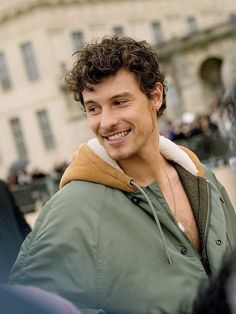 a man with curly hair smiling and wearing a green jacket in front of a building