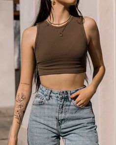 a woman with tattoos on her arms and chest wearing blue jeans, a brown crop top