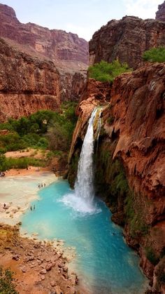 a waterfall in the middle of a canyon with people swimming under it and onlookers below