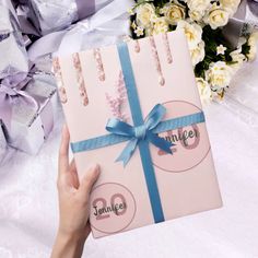 a person holding up a pink gift box with blue ribbon and flowers in the background