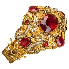 This exquisite bracelet, dating from the first half of the 20th century, showcases the remarkable craftsmanship of Austro-Hungarian jewelry. The wide band features vibrant, faceted garnet glass cabochons set in a luxurious gold gilt, accented by delicate faux seed pearls. Both the front and back of the bracelet display intricate openwork and pierced scroll designs, adding to its unique charm. The bracelet is equipped with a hidden, ornate tongue and box clasp, ensuring a secure fit while maintaining its elegant appearance. Although unmarked, the material is gold gilded metal, resembling lower grade silver or a metal mix, though it does not test positive for higher grade silver. Materials: Gold gilded metal Glass cabochons Faux seed pearls This stunning piece is perfect for collectors and t Hungarian Jewelry, Austro Hungarian, Bracelet Display, Box Clasp, Scroll Design, Seed Pearl, Gold Gilding, The 20th Century, Wide Bands