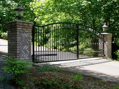 an iron gate and brick pillars in the middle of a driveway surrounded by greenery