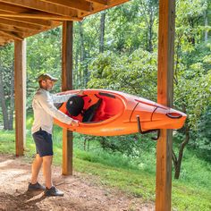 a man standing next to an orange kayak under a pergolated structure with trees in the background