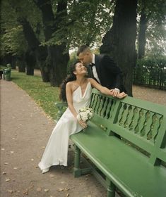 a bride and groom are sitting on a green bench in the park with their arms around each other