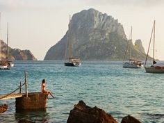 several sailboats in the water near a rocky island with a mountain in the background