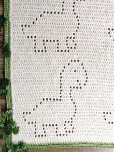 a white crocheted blanket with green trim on top of a wooden table next to a plant