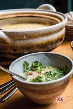 Rustic ceramic bowl of delicate broth garnished with fresh cilantro and green onions, served alongside a larger clay pot on a wooden table.