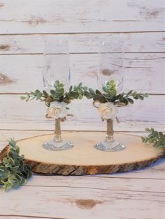 two wine glasses with flowers and greenery in them sitting on a piece of wood