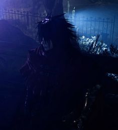 a man with black hair standing in front of a fence and some plants at night