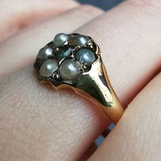 A delightful antique ring, featuring a central Emerald, surrounded by six white half-pearls in a traditional flower cluster shape. This would once have been a mourning ring, designed to hold a lock of hair of a departed loved one, as the compartment behind the head suggests.Unmarked, tested as 18ct gold.The head measures approximately 9.5mm in diameter.The approximate weight is 2.3 grams.Please note the antique ring box pictured is for display purposes only and is not included in this sale. Than Antique Ring Box, Traditional Flower, Lock Of Hair, Gold Pearl Ring, Gold Cushions, Flower Cluster, Antique Ring, White Band, Small Rings