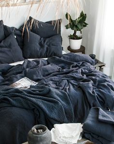 an unmade bed with dark blue sheets and blankets on it, next to a potted plant