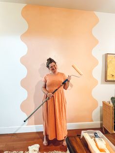 Young lady wearing a peach colored mid calf dress is holding a paint roller with an extender. She is standing in front of a peach colored squiggle painted on a white wall to create an accent and feature wall in her apartment Wall Painting Ideas Creative, Long Hall, Plain Wall, Room Unique, Diy Wall Painting, Accent Wall Paint