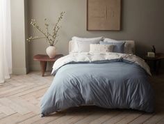 a bed with blue sheets and pillows in a room next to a window, vase with flowers