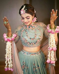 a woman in a blue and pink lehenga with flowers on her head, holding two hands up