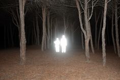 two people standing in the middle of a forest at night with light coming from their faces