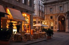 an outdoor cafe with tables and chairs on the sidewalk in front of buildings at night