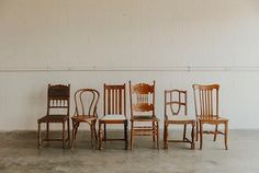 six wooden chairs are lined up against a white wall in an empty room with concrete flooring