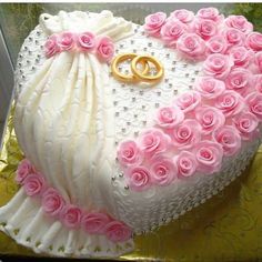 a heart shaped cake decorated with pink roses and two gold wedding rings on the top