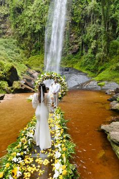 a bride and groom standing in front of a waterfall with flowers on the ground next to it