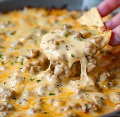 a hand holding a tortilla chip over a casserole dish with cheese and ground beef