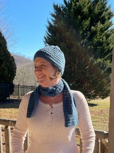 a woman wearing a knitted hat and scarf standing on a porch with trees in the background