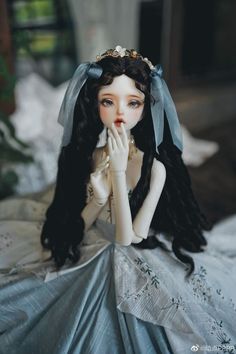 a doll with long black hair wearing a blue dress sitting on top of a bed