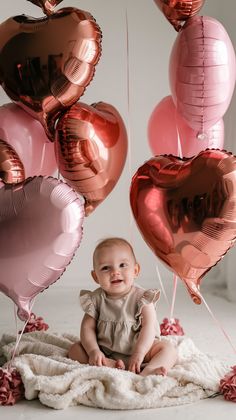 a baby sitting on a blanket surrounded by pink and red heart - shaped helium balloons