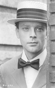 a man in a suit and bow tie standing next to a brick wall wearing a hat