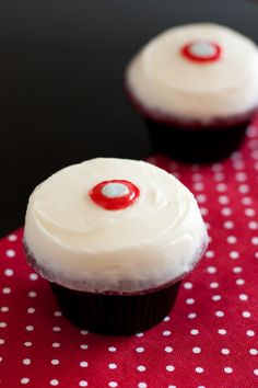 two cupcakes with white frosting and a red eye on top sitting on a table