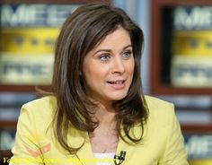 a woman in a yellow blazer sitting at a table with her hand on her chest