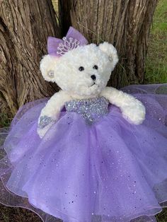 a white teddy bear wearing a purple dress sitting next to a tree in the grass