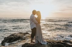 a man and woman are standing on rocks by the ocean at sunset with their arms around each other