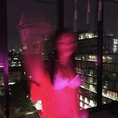 a woman standing in front of a window with the city lights lit up behind her