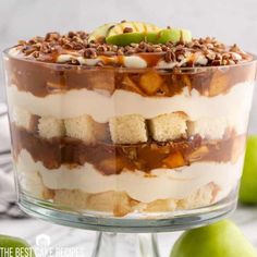 a layered dessert with apples and nuts in the middle on a glass cake stand next to two green apples