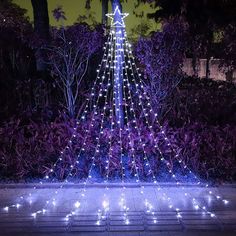 a lighted christmas tree in the middle of a garden with purple bushes and trees around it